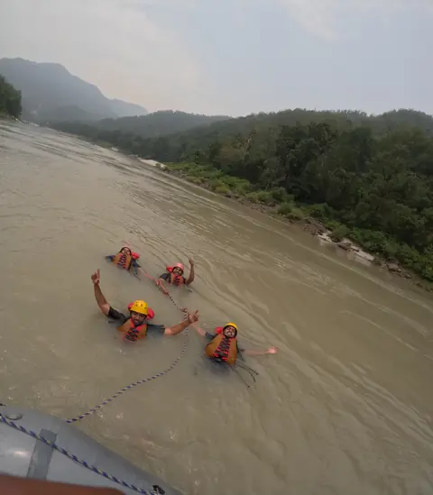 Four rafters hold the raft’s safety rope in the river and wave at the camera