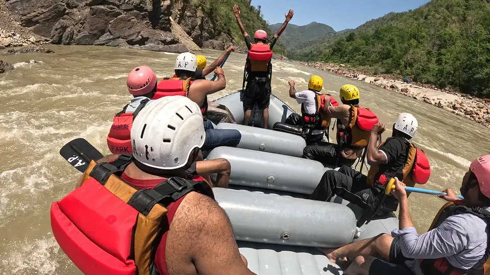 The guide stands at the front of an Adventure Park raft with arms raised as the crew paddles on